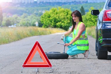 Yedek tekerlek değişimi ile kız umutsuzluk. Araba sürerken yolda delikli tekerlek. Yansıtıcı yelekli kız tekerlek anahtarını tutar ve yedek tekerleği yuvarlar. Önünde acil durdurma işareti ayarlandı. 