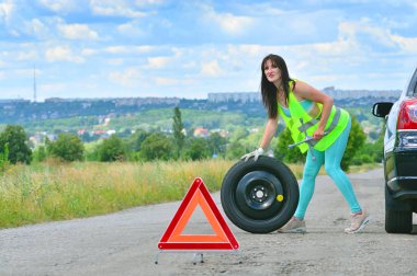 Yedek tekerlek değişimi için yardım uyamasını uman bir kız. Araba sürerken yolda delikli tekerlek. Yansıtıcı yelekli kız tekerlek anahtarını tutar ve yedek tekerleği yuvarlar. Önünde acil durdurma işareti ayarlandı. 