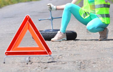 Yedek tekerlek değişimi. Yansıtıcı yelekli kız, tekerlek anahtarını ve yedek tekerleği tutuyor. Önünde acil durdurma işareti ayarlandı. 