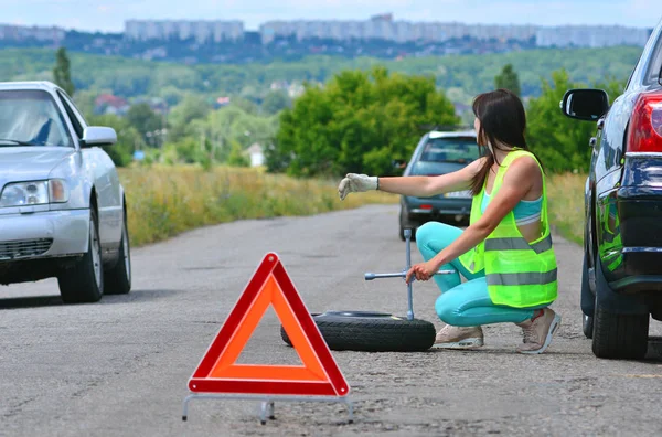 Girl hoping for help with spare wheel replacement. Punched wheel on the road while driving. Girl l in reflective vest holds wheel wrench and rolls the spare wheel. Emergency stop sign set in front.