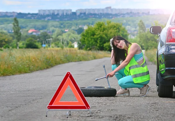 Girl despair with spare wheel replacement. Punched wheel on the road while driving. Girl l in reflective vest holds wheel wrench and rolls the spare wheel. Emergency stop sign set in front.