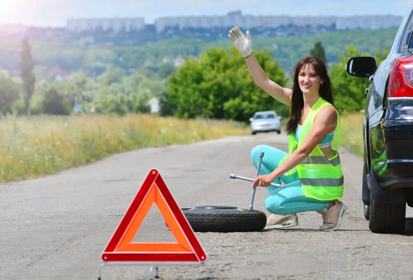 Girl searching for help with spare wheel replacement. Punched wheel on the road while driving. Girl l in reflective vest holds wheel wrench and rolls the spare wheel. Emergency stop sign set in front.