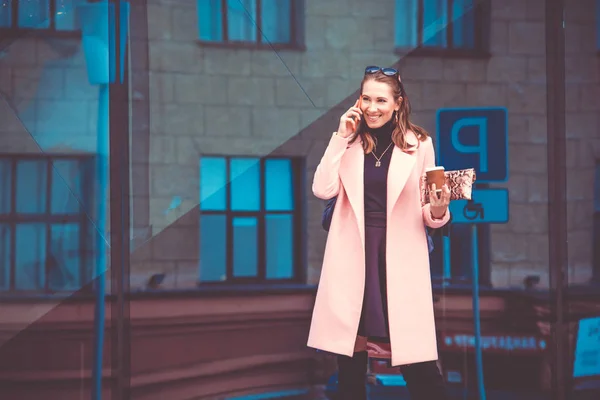 Business lady on a coffee break in a lunch time. She is talking on the phone and laughing. In his other hand holds a paper cup of coffee to go.