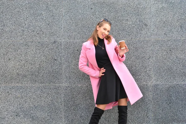 Beautiful and happy woman in pink coat  and knee-high boots standing with coffee cup and posing on camera on granite wall background