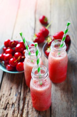 Kırmızı böğürtlen ve meyvelerden yapılmış kırmızı smoothie ile yapılmış cam şişeler. Yakınlarda kiraz ve çilekli tabak var. Dikey bileşim. 