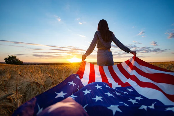 Young happy girl with the flag of America at the wheat field against the sunset