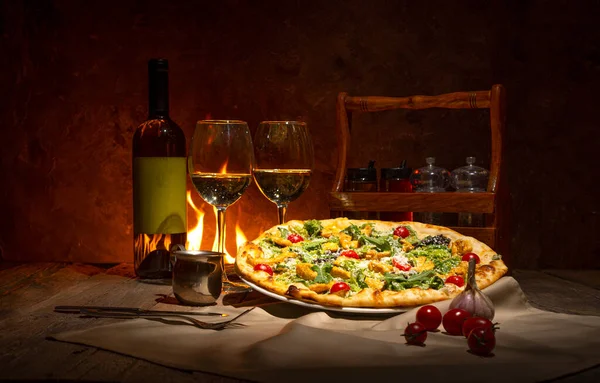 Pizza, bottle of white wine and two wine glasses  against the fireplace. Evening romantic mood in italian restaurant.