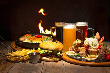 Bira partisi masası arka planında bira bardakları ve farklı yemekler. Hamburger, kızarmış sosis, patates kızartması ve masada ızgara et. Arkaplanda yangın alevi. 