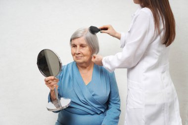 Yaşlı bir kadının saçını tarayan bir sağlık görevlisi. Yuvarlak aynaya bakan yaşlı kadın.. 