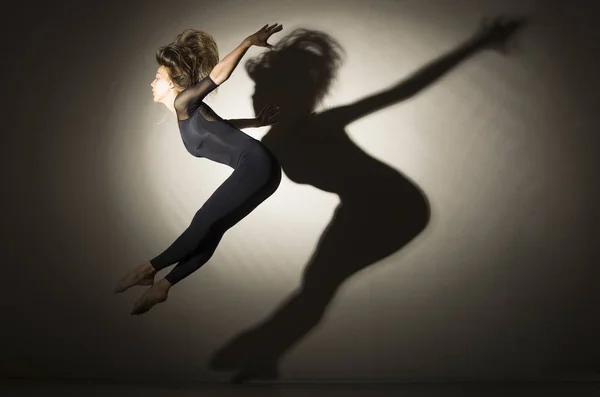 Девушка в темноте совершает гимнастический прыжок, на белом фоне появляется тень от формы . — стоковое фото