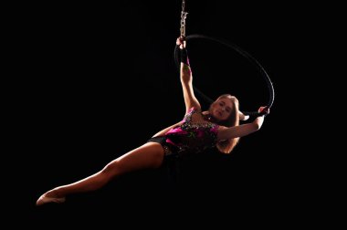 Koyu renk bir takım elbiseyle hava akrobasisi yapan genç bir kadın çevre aydınlatması altında bir hava halkası içinde egzersiz yapıyor. Karanlık bir arkaplanda stüdyo çekimi.