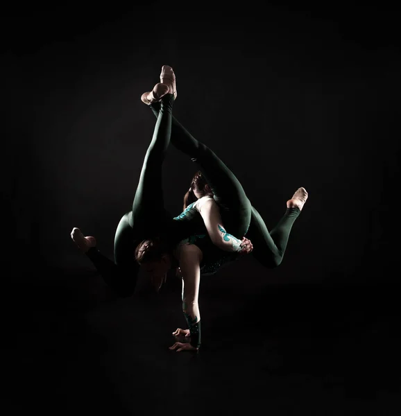 Two young female equilibrists perform acrobatic elements on a dark background in contoured lighting. — Stock Photo, Image