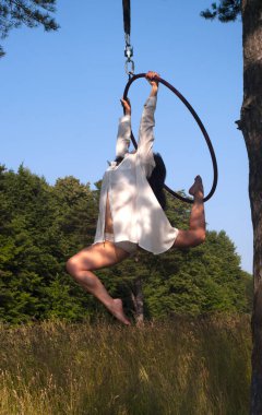 Beyaz sahne kostümlü bir kız açık havada havadaki bir hava halkası içinde egzersiz yapıyor, ormanın ve gökyüzünün arka planına karşı. Sokakta fotoğrafçılık.