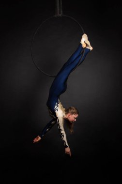 Uzun saçlı, mavi ve beyaz takım elbiseli, ince bir kız-hava akrobatı, hava ringinde egzersiz yapıyor. Karanlık bir arkaplanda stüdyo çekimi.