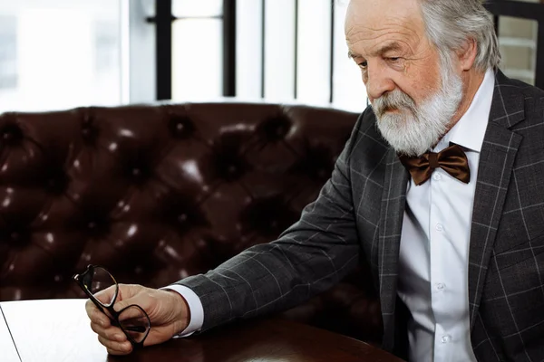 Unhappy old person holding his glasses and sitting on the red couch — Stock Photo, Image