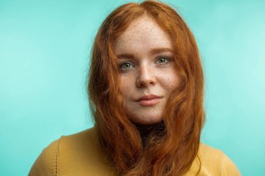 Kıvırcık saçlı kızıl kız mavi arka plandaki kameraya bakarak gülümsedi.