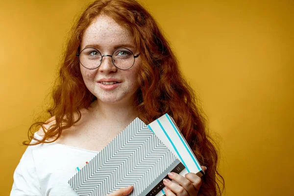 Rousse jeune femme tenant des livres vierges regardant la caméra isolée jaune — Photo