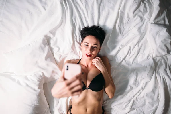 Attractive sexy brunette woman lies on bed in lingerie and taking selfie on phone. — Stock Photo, Image