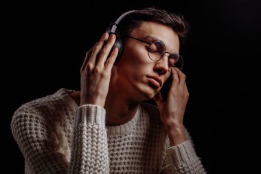Gözlüklü ve kulaklıklı genç adam siyah arka planda müzik dinliyor.