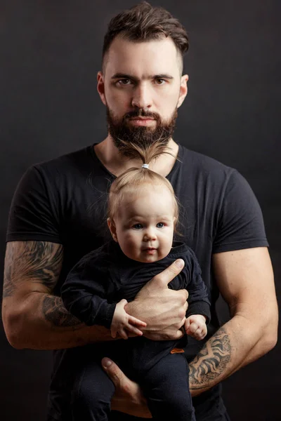 Knappe getatoeëerd beared man houdt van schattige kleine baby op zwarte achtergrond — Stockfoto