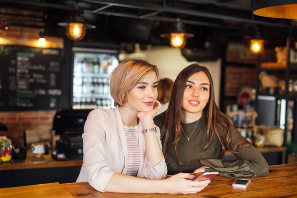Dos atractivas amigas jóvenes charlando en el teléfono inteligente en la cafetería — Foto de Stock