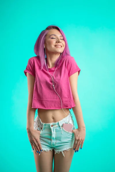 Nieśmiała Pani z różowymi włosami pozowanie na niebieskim tle słuchania muzyki w słuchawkach. — Zdjęcie stockowe