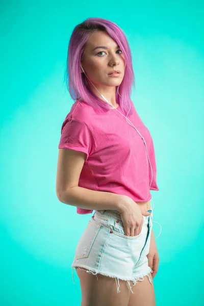 Ντροπαλή κοπέλα με ροζ μαλλιά ποζάρει πάνω από μπλε φόντο ακούγοντας μουσική στα ακουστικά. — Φωτογραφία Αρχείου