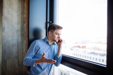 Ofisin penceresinde duran akıllı telefonu kullanan kendine güvenen genç işadamı.