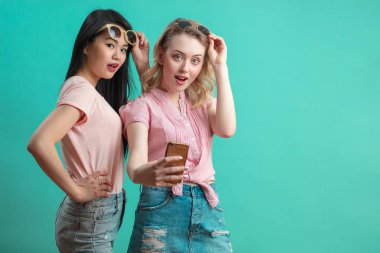 Mutlu genç kızlar mavi duvara karşı akıllı telefonlarıyla selfie çekiyorlar.