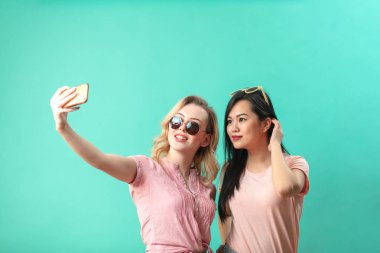 Mutlu genç kızlar mavi duvara karşı akıllı telefonlarıyla selfie çekiyorlar.