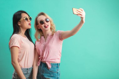 Mutlu genç kızlar mavi duvara karşı akıllı telefonlarıyla selfie çekiyorlar.