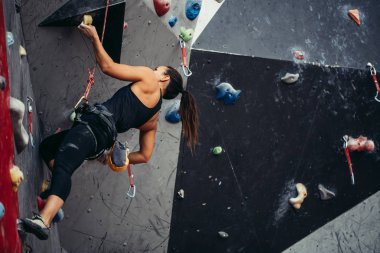 Siyah kıyafetli profesyonel dağcı kadın, kayalık spor salonunda çalışırken yapay kaya duvarının tepesine ulaşıyor. Düşük açılı dikey hava kaslı kadın vücudu üzerine gerilme görüntüsü.