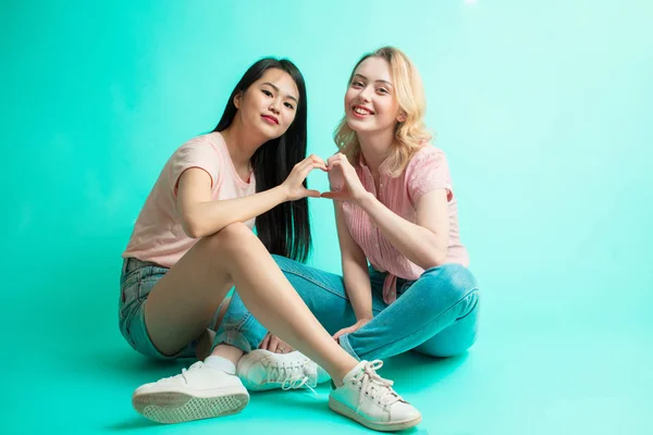 Chicas jóvenes mostrando el signo del corazón con las manos sentadas sobre fondo azul — Foto de Stock