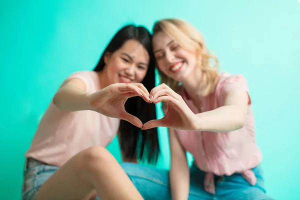 Chicas jóvenes mostrando el signo del corazón con las manos sentadas sobre fondo azul — Foto de Stock