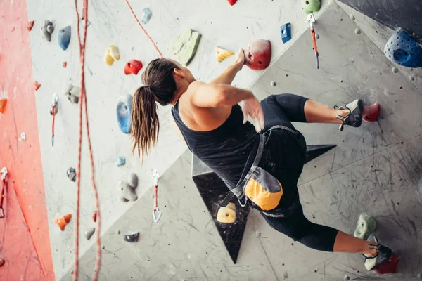 Beautiful young woman in black outfit climbing on practical wall in gym,  bouldering, extreme sport, rock-climbing concept. Stock Photo