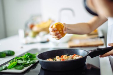 Genç ev hanımı mutfakta yemek pişiriyor, tavaya portakal sıkıyor.