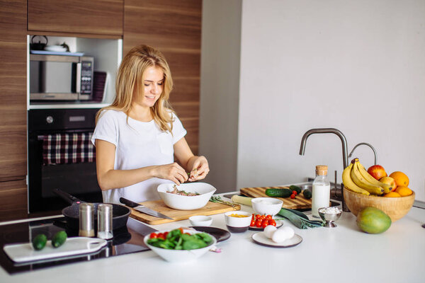 Портрет счастливой жены в белой футболке, готовящей ужин на кухне