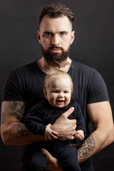 Красивий татуйований бородатий чоловік тримає милу дитину на чорному тлі — стокове фото