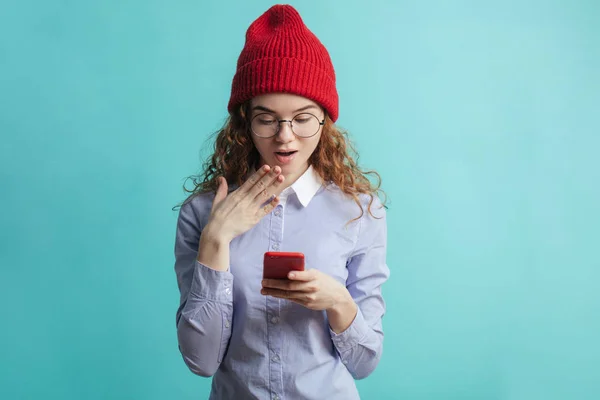 Удивлённая девушка в очках, красная кепка. голубая рубашка смотрит на смартфон — стоковое фото