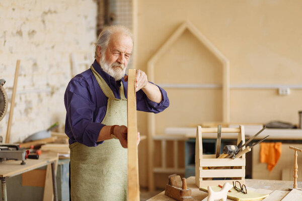 close up photo of working senior citizen with closed one eye holding plank on hands and choosing planks for furniture