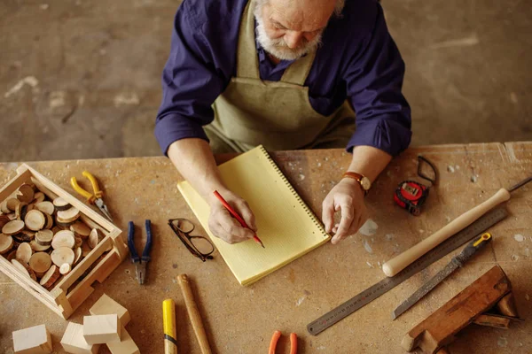 old carpenter wearing working clothes sitting at wooden desk and making notes