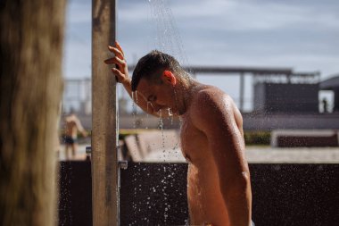 Yakışıklı, üstsüz, beyaz bir adam sahilde duş alıyor..