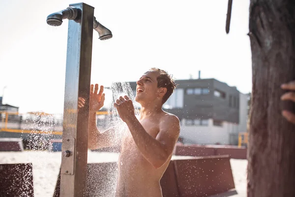 Красивый молодой топлесс белый мужчина принимает душ на открытом воздухе на пляже. — стоковое фото