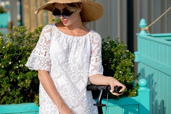 Portret van Trendy vrouw, mooi blond meisje in witte jurk, zonnebril en hoed paardrijden scooter — Stockfoto