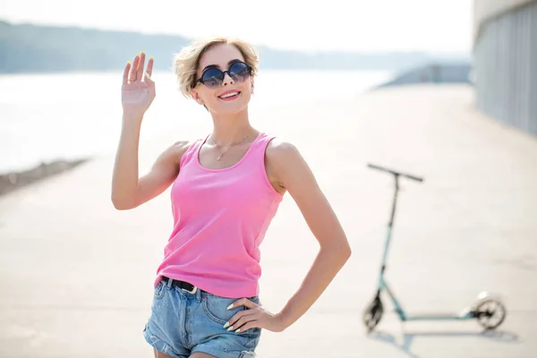 Chica impresionante en la calle de la ciudad en patinete scooter. Disfrutando de un día soleado. Usar gafas de sol, pantalones cortos — Foto de Stock