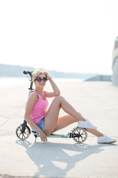 Mulher loira positiva sentada na scooter pontapé azul — Fotografia de Stock