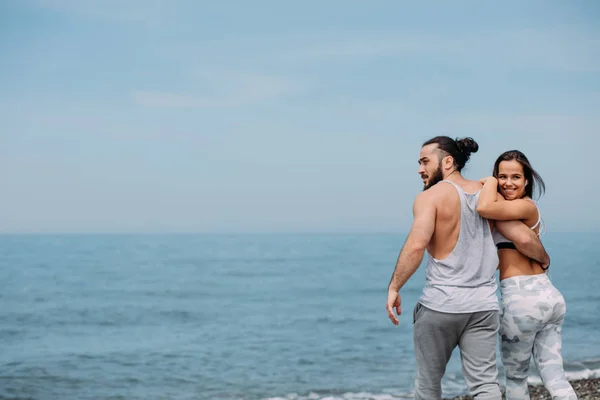 Joven pareja de fitness abrazándose de pie en la playa, mirando al horizonte —  Fotos de Stock