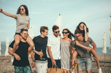 Bir grup arkadaş sahilde yürüyor, eğleniyor, kadınlar erkekleri sırtında taşıyor, komik bir tatil.
