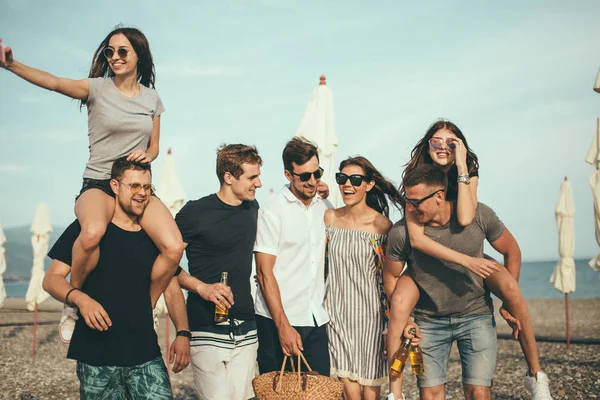 Grupo de amigos caminando en la playa, divertirse, las mujeres a cuestas en los hombres, vacaciones divertidas —  Fotos de Stock