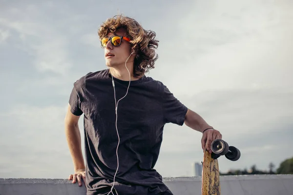 Hipster man die in de buurt van betonnen wand met longboard met behulp van zijn telefoon en de hoofdtelefoon — Stockfoto